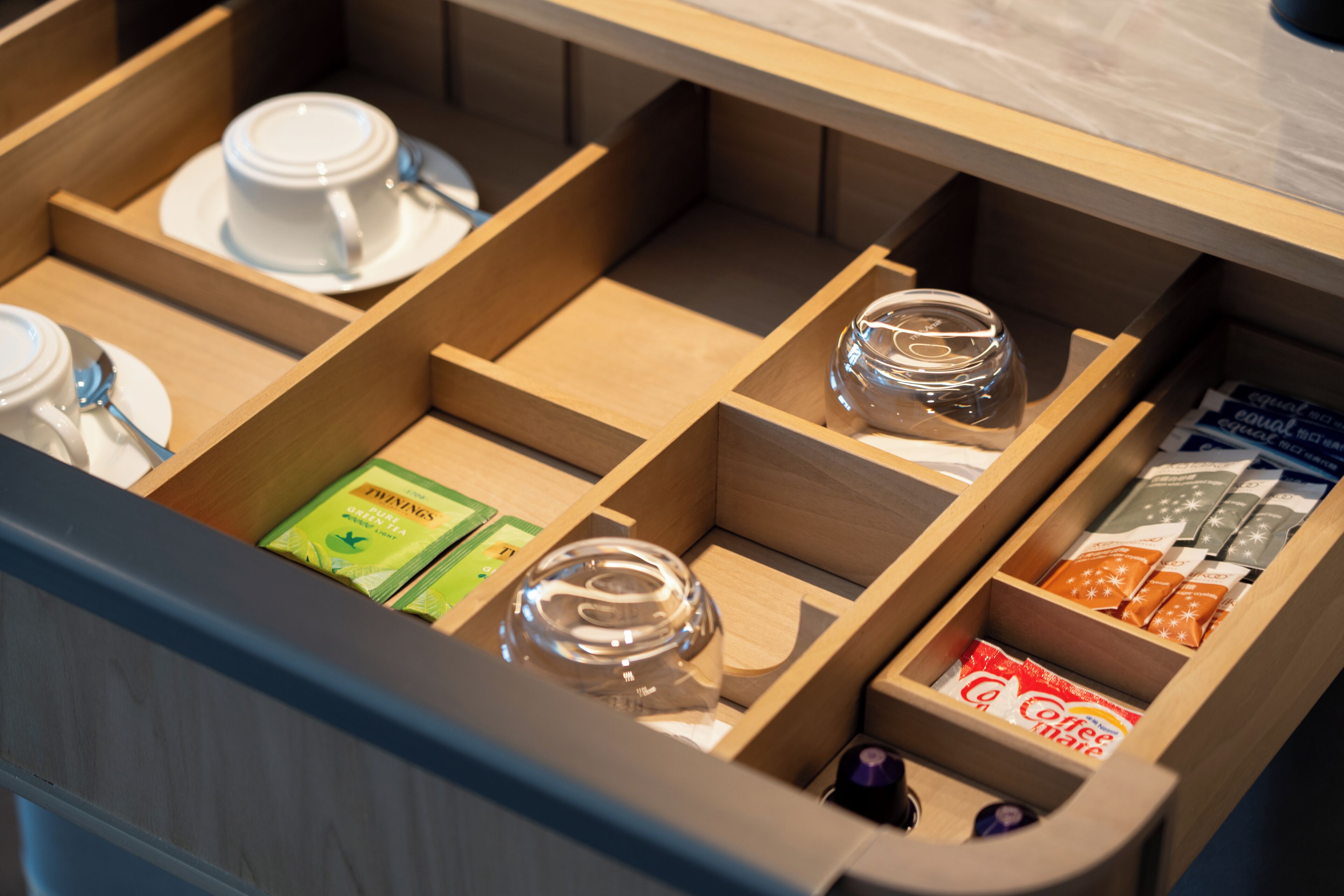 A drawer with compartments containing cups, saucers, a glass, tea bags, and coffee packets.