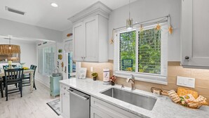 A modern kitchen with white cabinetry, a double sink, and a window with a view of greenery. - Peaceful Holmes by Beach Boutique Rentals (Holmes Beach)