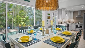 A dining area with a table set for four, featuring yellow plates and blue glassware, a woven pendant light, and a view of the outdoors through large windows. - Peaceful Holmes by Beach Boutique Rentals (Holmes Beach)