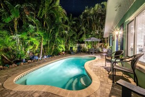 Une piscine entourée d’une végétation luxuriante, avec un espace patio comprenant des chaises et une table.