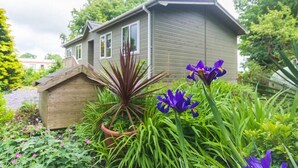 Una casa con techo inclinado, un cobertizo de madera y un jardín con flores moradas y plantas verdes.