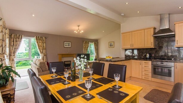 Comedor con mesa de madera dispuesta para seis personas, rodeada de sillas. Se observa una cocina con electrodomésticos modernos y armarios de madera.