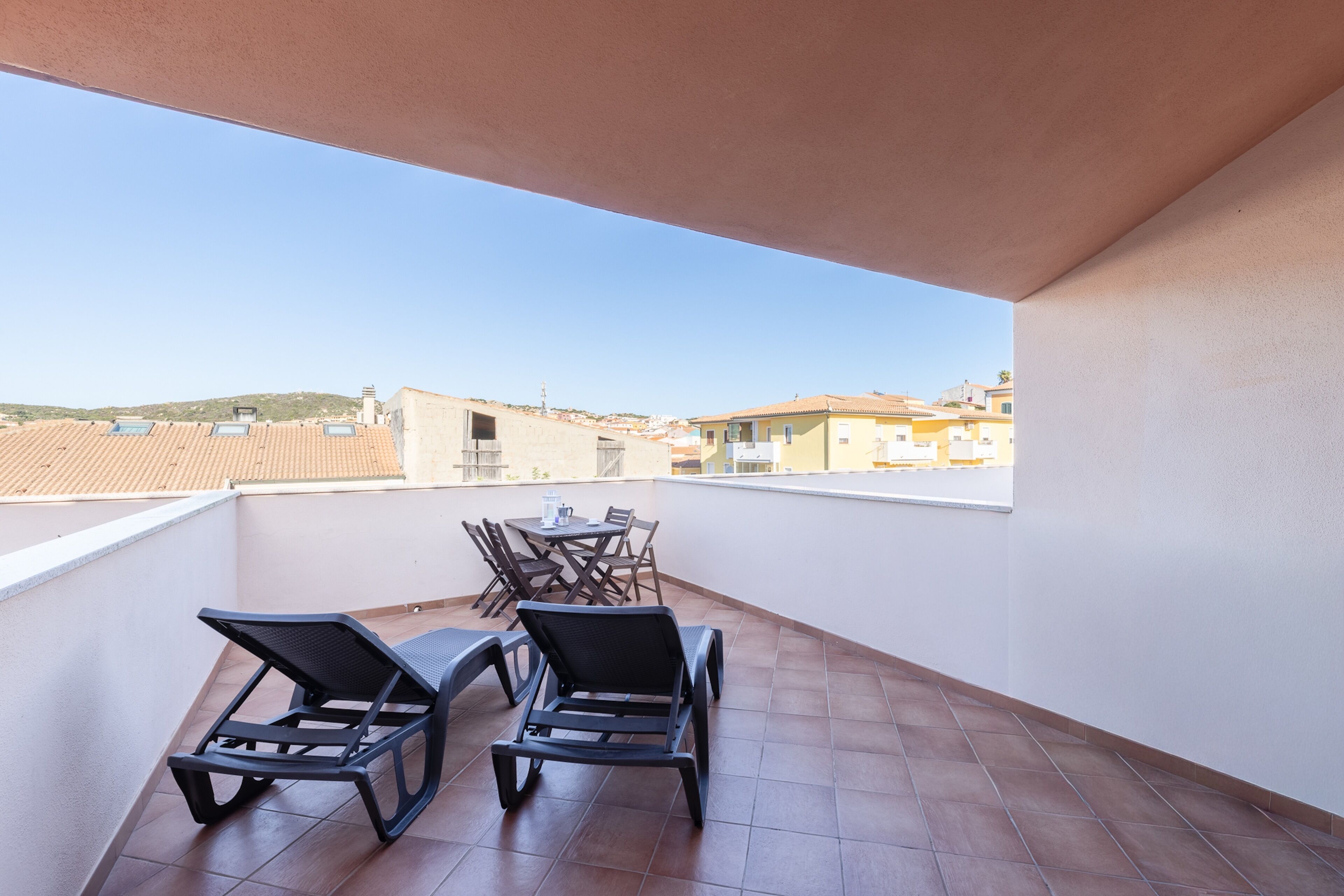 A balcony with two lounge chairs and a table, overlooking a residential area with tiled roofs.