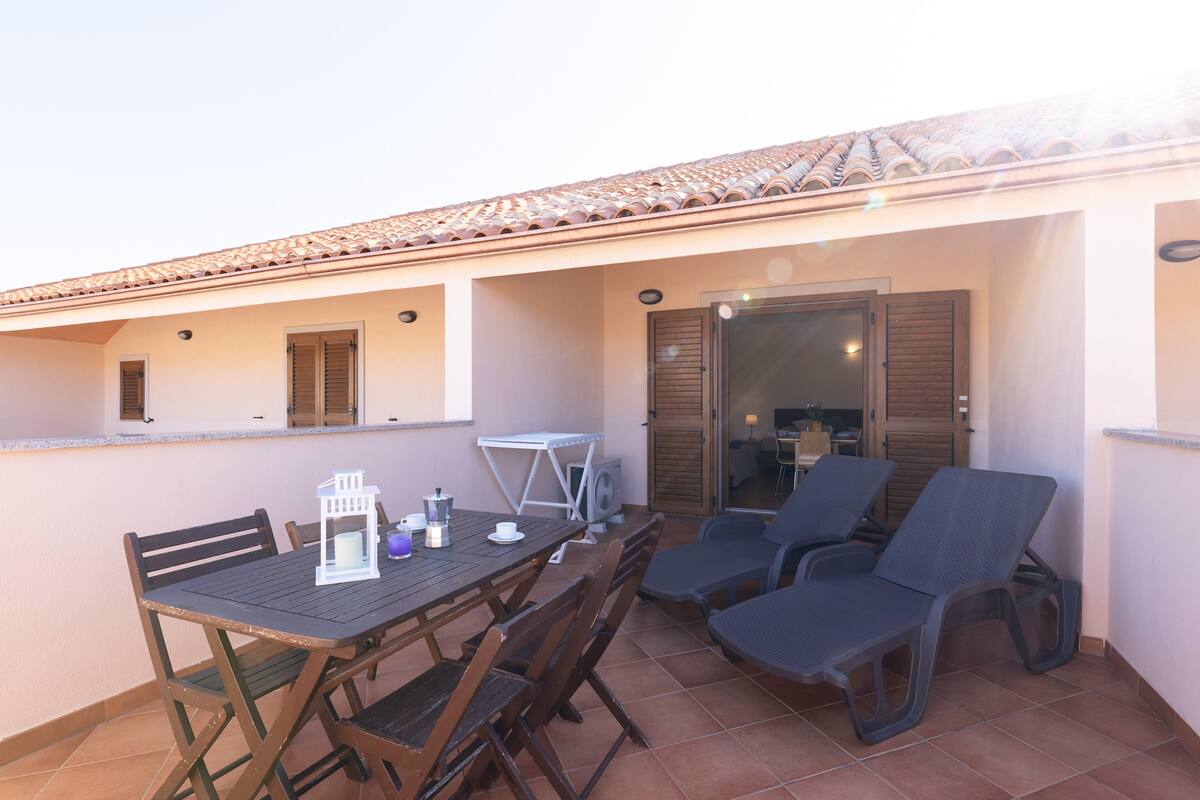Eine Terrasse mit Tisch und Stühlen, ein kleiner Tisch mit einer Laterne und eine Tür, die in einen Innenraum führt.
