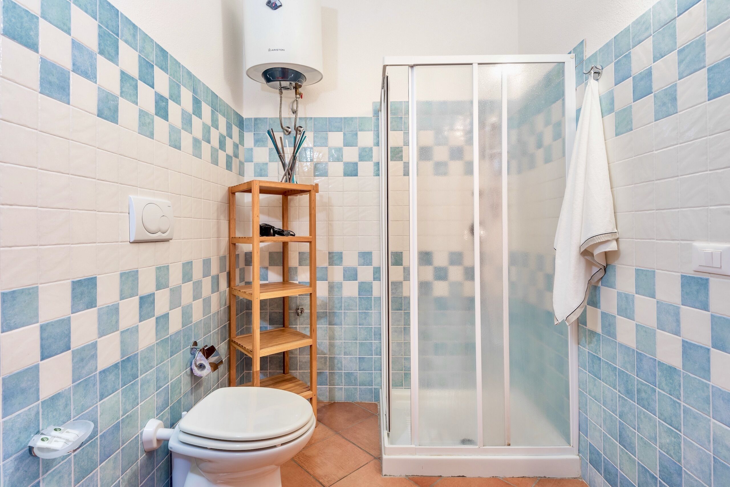 A bathroom with a toilet, a wooden shelf, a glass shower enclosure, and a towel hanging on the wall.
