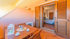 A wooden dining table with a white lantern, a cup, and a small house model. There is a wooden door with shutters and a view into a room with a bed and a window. - Residence con piscina a 300 mt dal mare (Santa Teresa di Gallura)