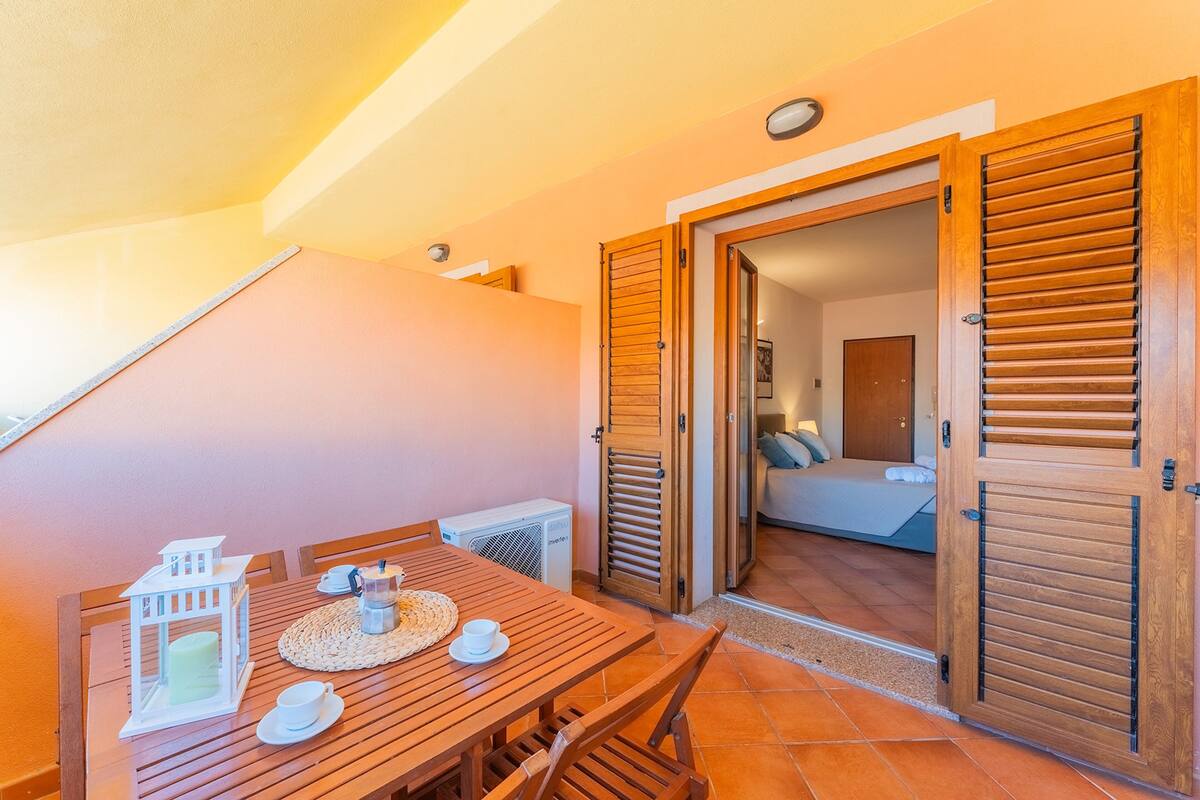 Ein Holzesstisch mit einer weißen Laterne, einer Tasse und einem kleinen Hausmodell. Eine Holztür mit Fensterläden und Blick in einen Raum mit Bett und Fenster.