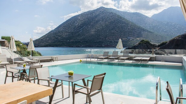 Área junto à piscina com espreguiçadeiras e mesas, com vista para uma paisagem montanhosa e um corpo d'água.