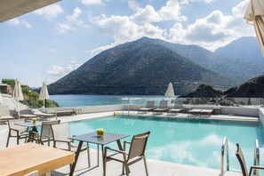 A poolside area with lounge chairs and tables, overlooking a mountainous landscape and a body of water. - Bali Diamond Adults Only - by Checkin (Mylopotamos)