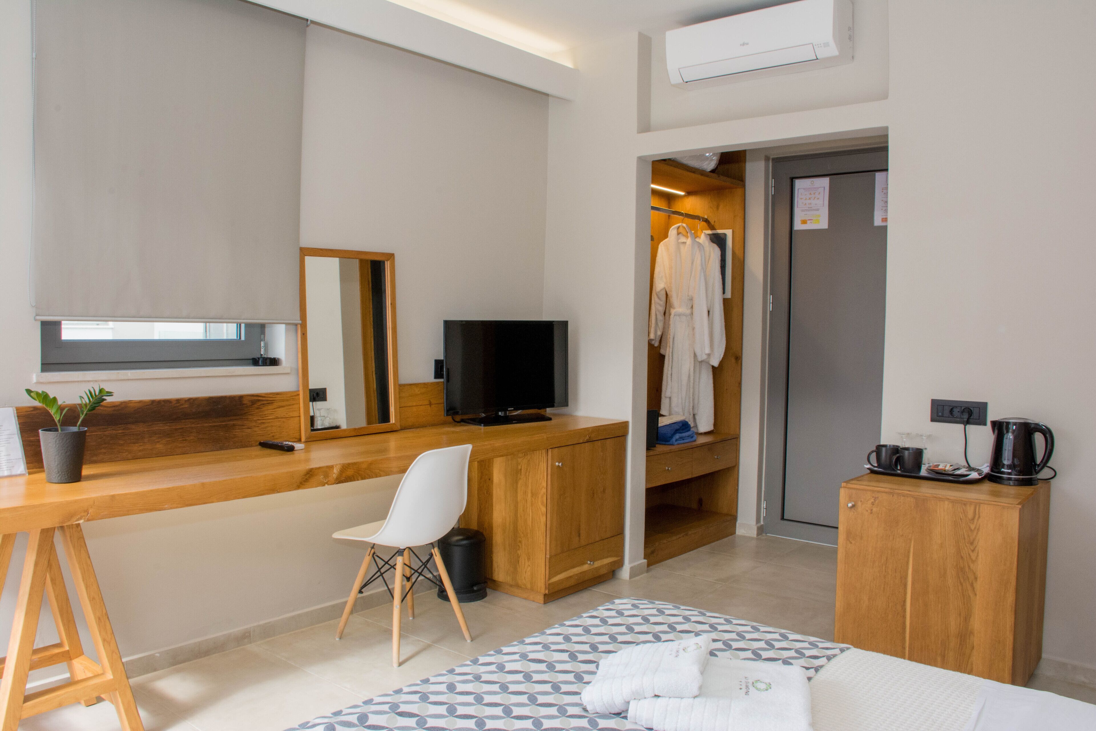 A hotel room with a wooden desk, a television, a chair, a bed with a patterned bedspread, a small potted plant, and a wardrobe with hanging clothes.