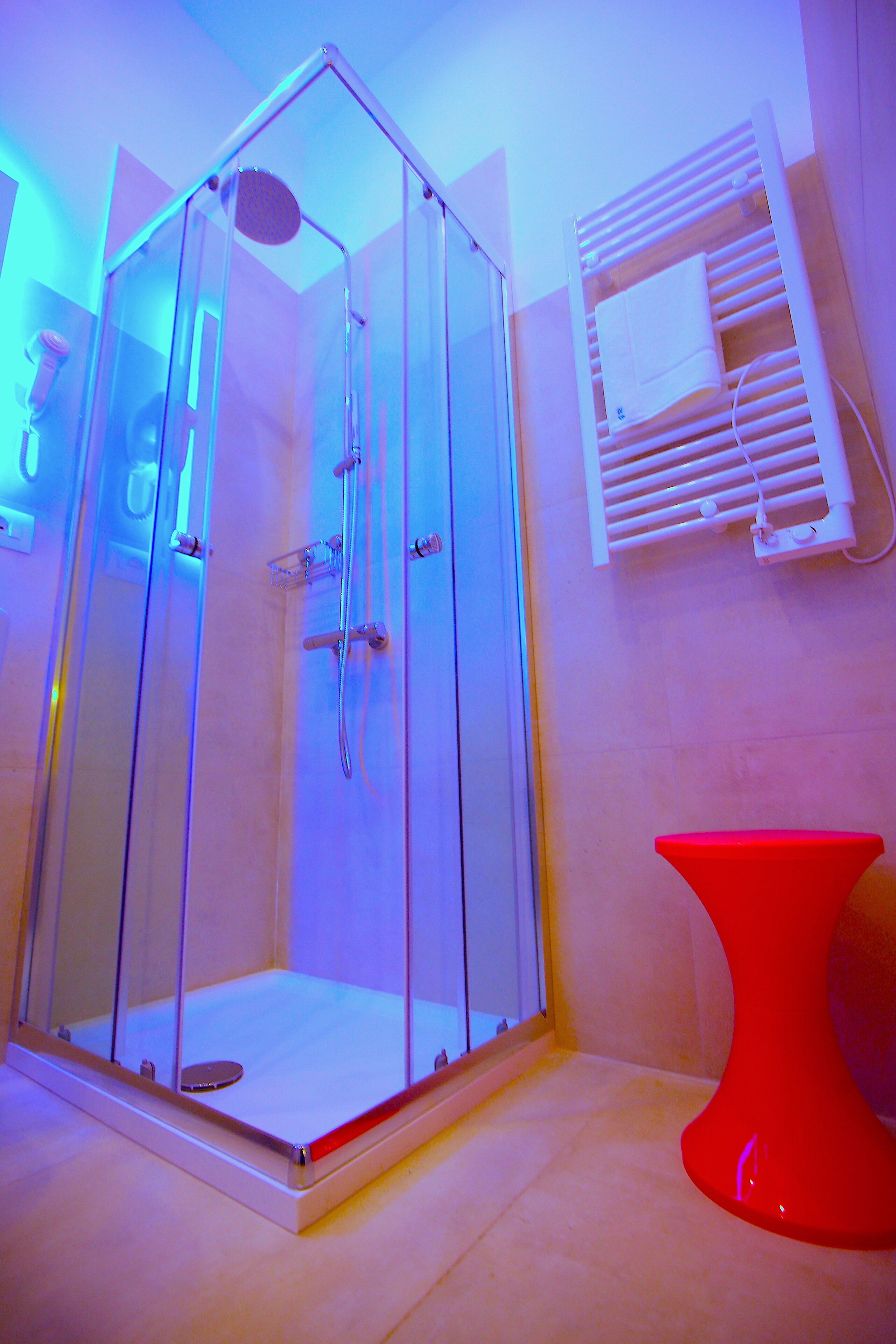 A glass-enclosed shower with a red accent piece, a white towel rack, and a red stool.