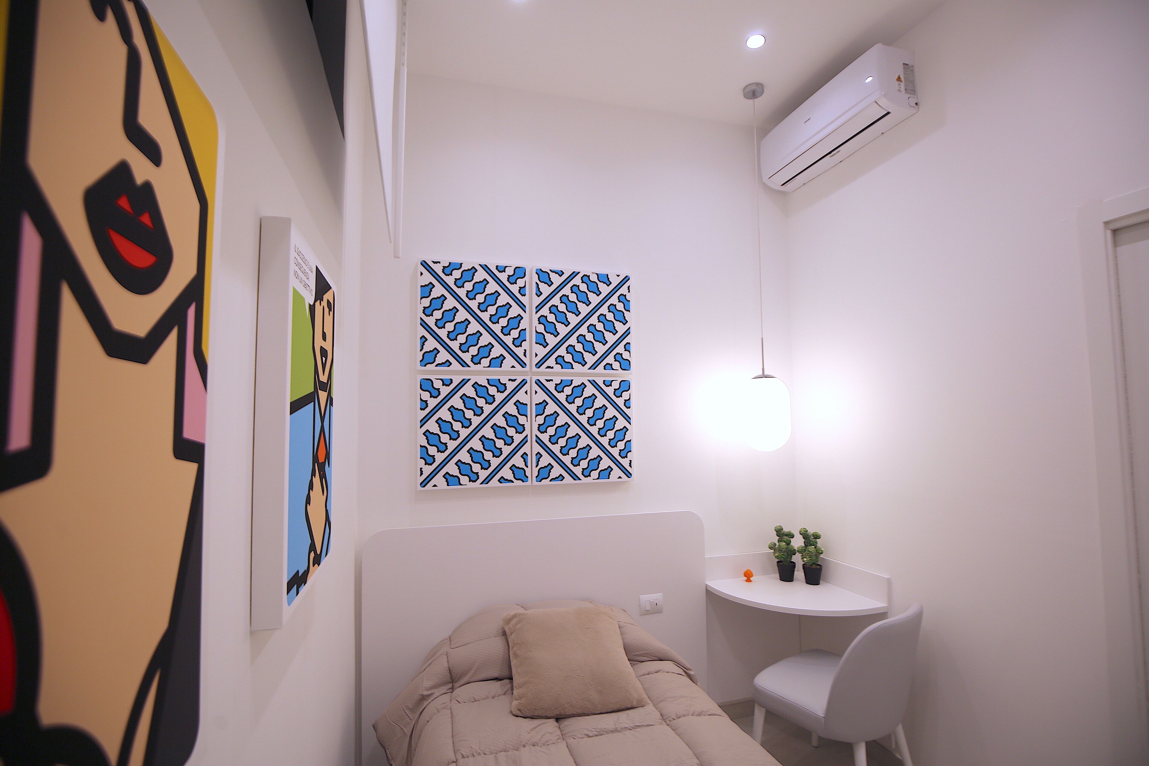 Una habitación pequeña y moderna con una cama, un escritorio y una silla. Hay un equipo de aire acondicionado en la pared y una obra de arte decorativa sobre la cama.
