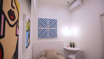 Una habitación pequeña y moderna con una cama, un escritorio y una silla. Hay un equipo de aire acondicionado en la pared y una obra de arte decorativa sobre la cama.