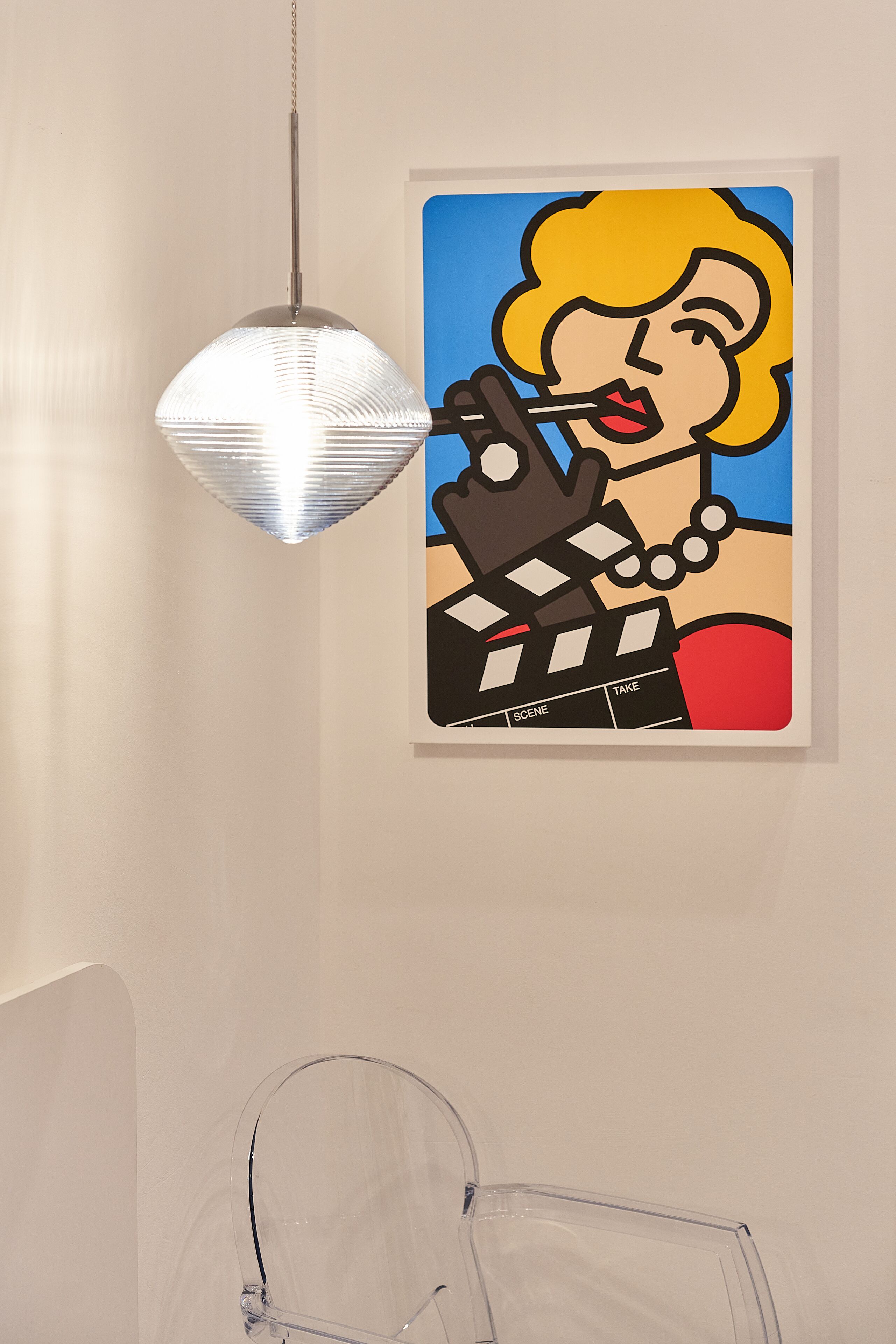 Een transparante glazen stoel, een hanglamp en een ingelijst pop-art kunstwerk aan een muur.