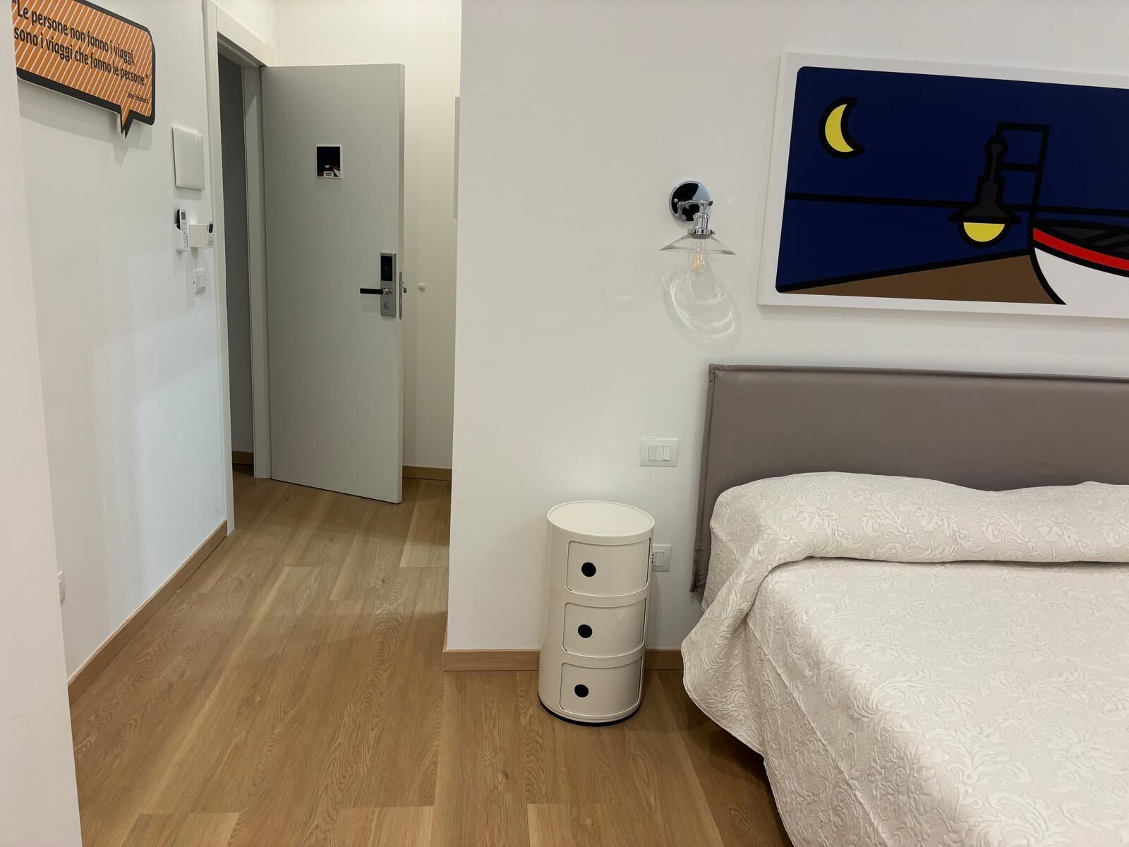Une chambre à coucher comprenant un lit, une table de chevet, une lampe fixée au mur, une porte donnant sur une autre pièce et une petite armoire blanche.