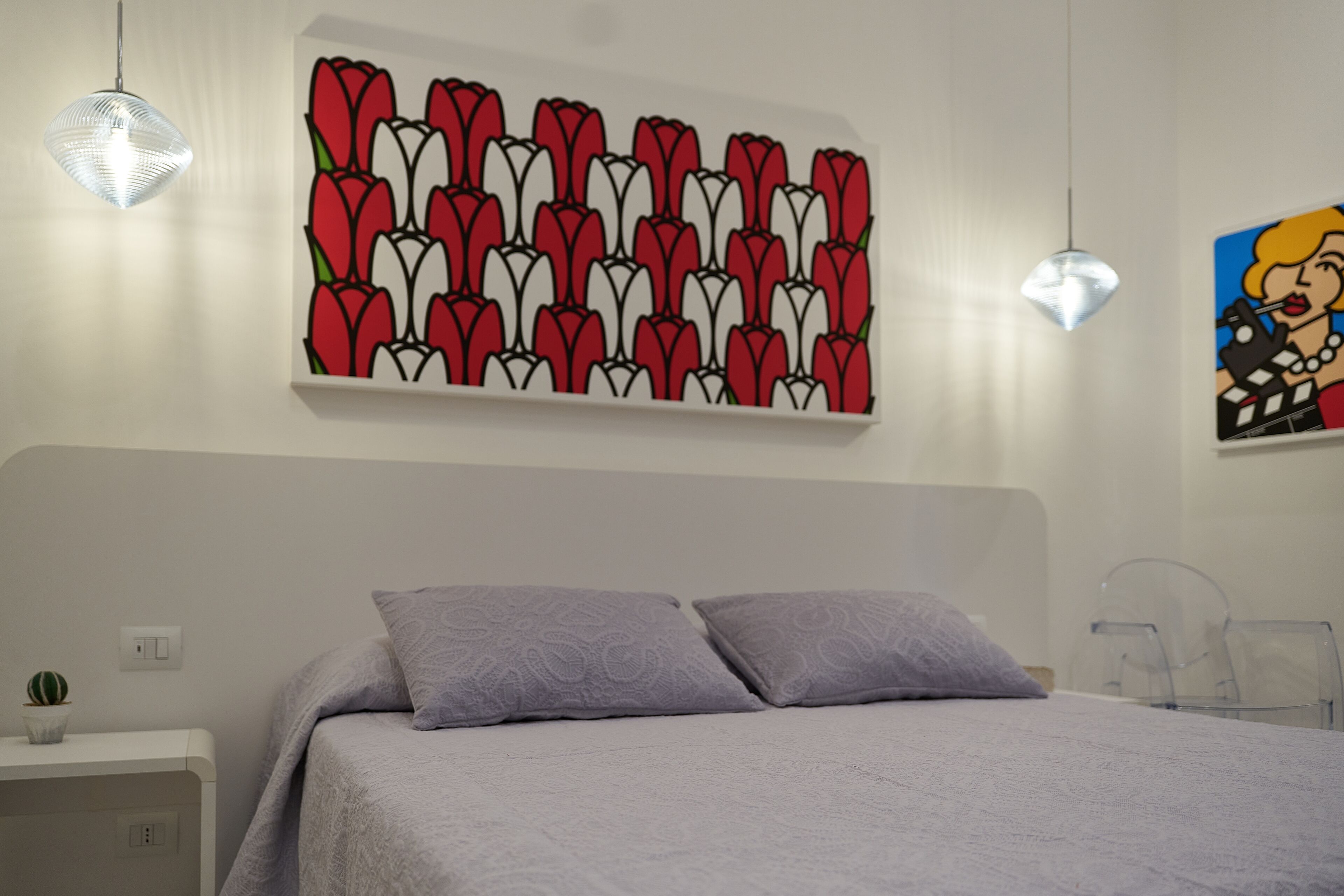 A bedroom with a bed, two pillows, a wall-mounted artwork of red and white flowers, and a pendant light.