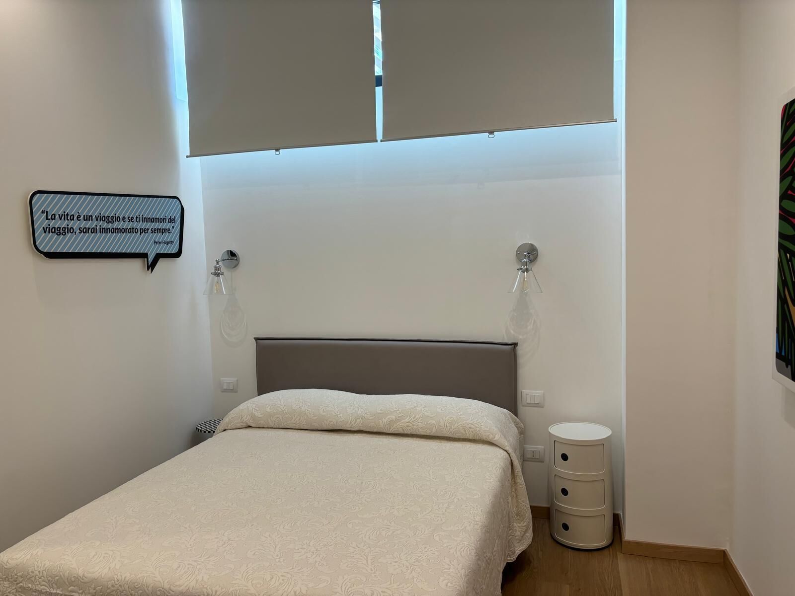 A small, neatly arranged bedroom with a bed, bedside lamps, a wall-mounted quote, and a small bedside table.