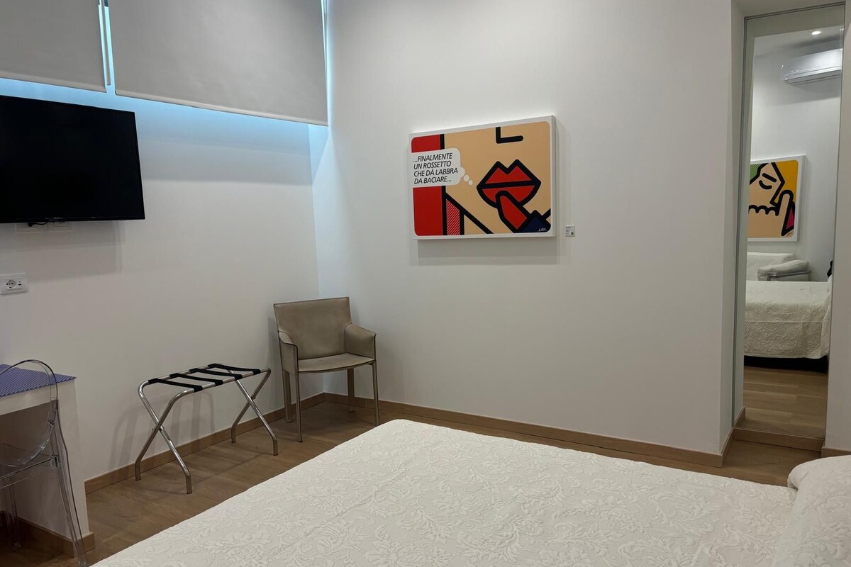 A hotel room with a bed, a chair, a small table with a towel, a wall-mounted TV, and a painting.