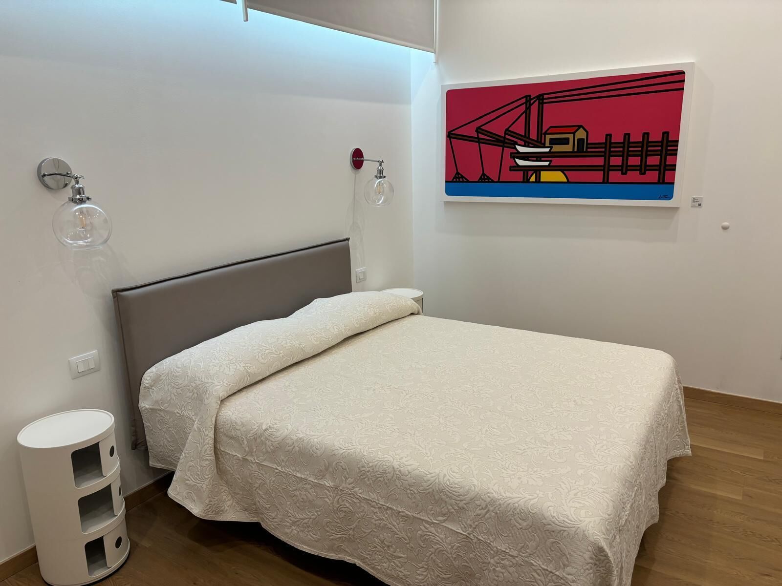 A modern bedroom with a bed, a wall-mounted lamp, a small bedside table, and a painting.