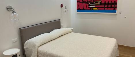 A modern bedroom with a bed, a wall-mounted lamp, a small bedside table, and a painting.