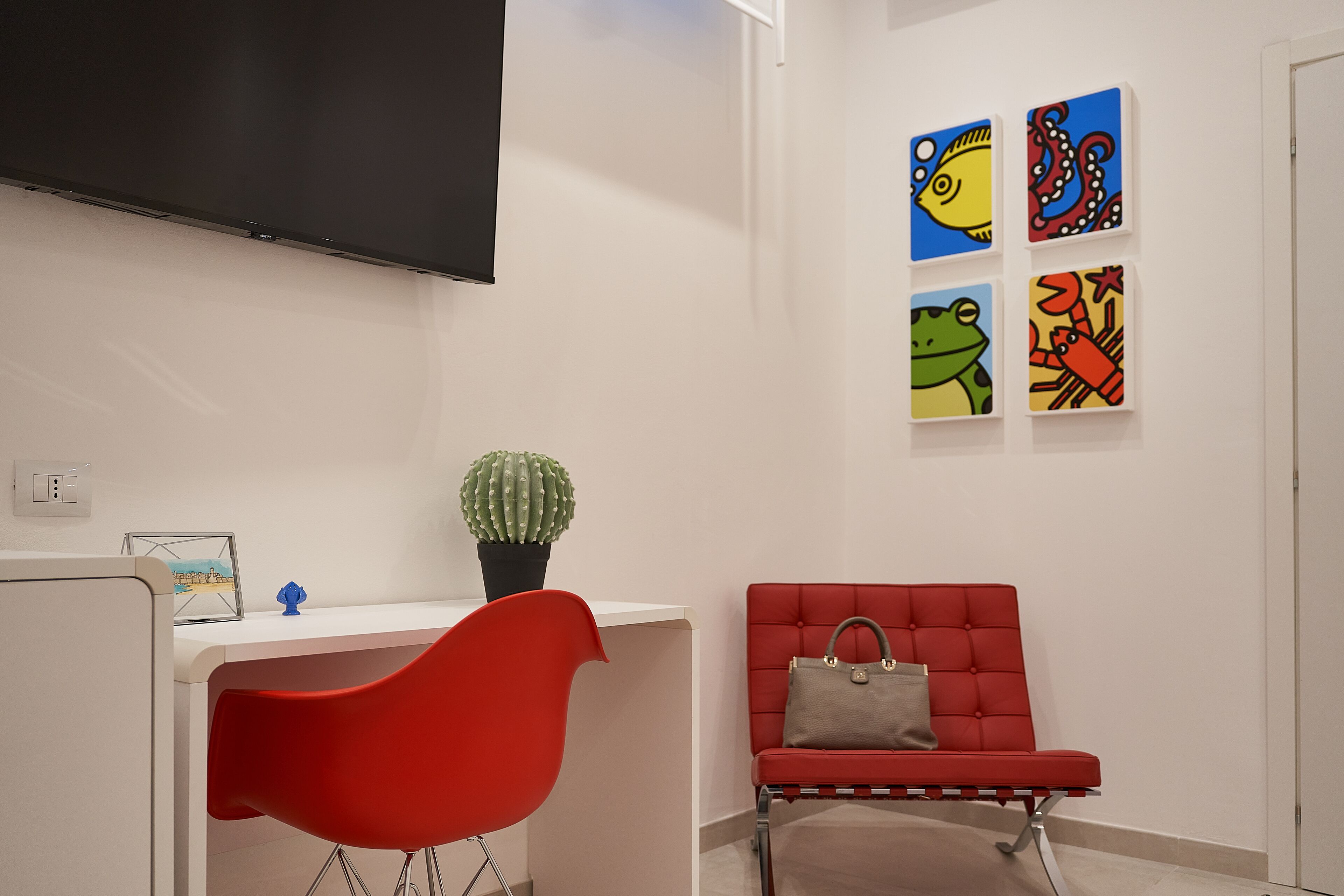 Quarto moderno com escrivaninha branca, cadeira vermelha, sofá vermelho com uma bolsa e quatro quadros coloridos na parede.