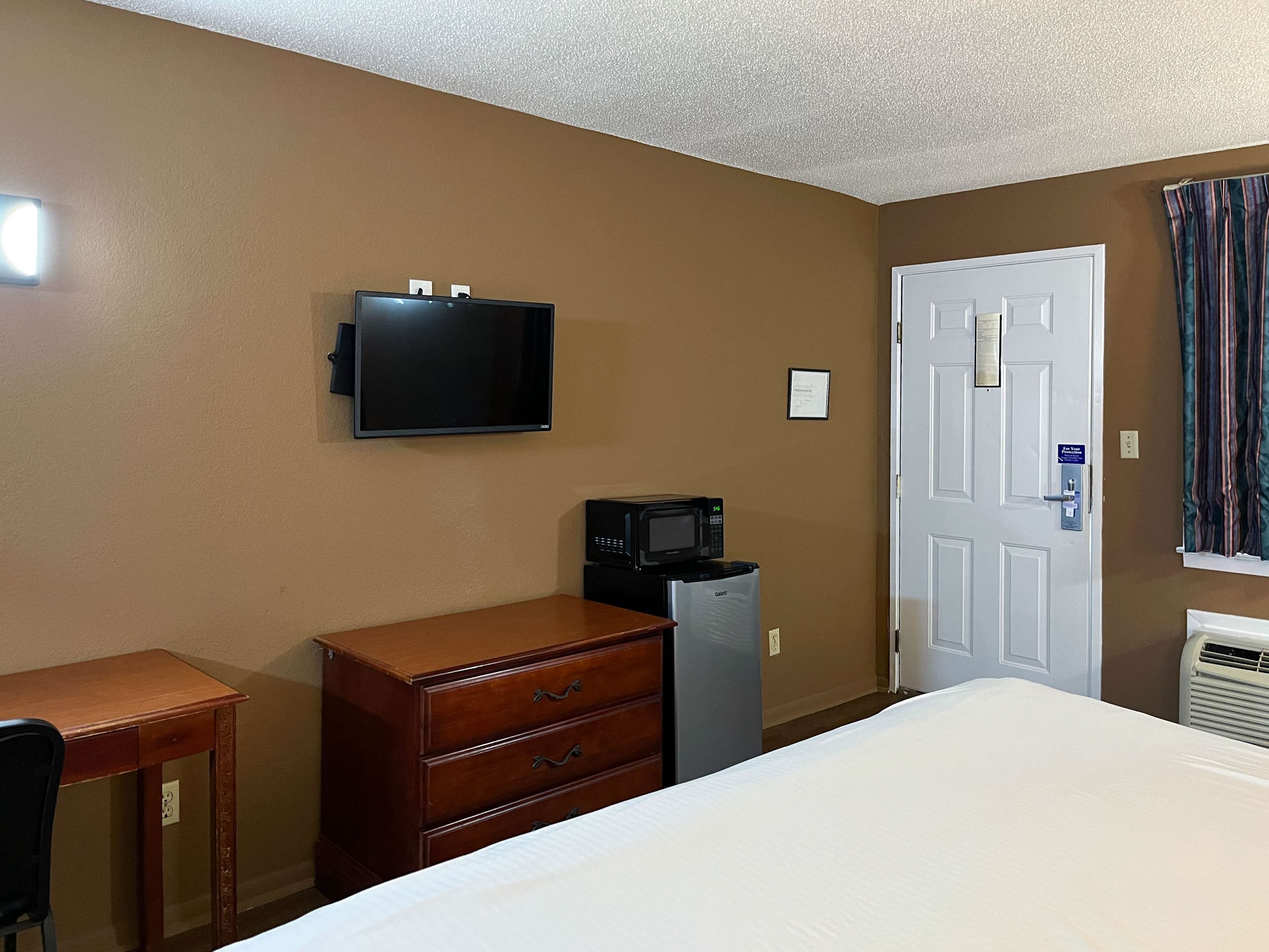 Habitación de hotel con cama, cómoda, escritorio, televisor, microondas, refrigerador pequeño y una puerta con un cartel.