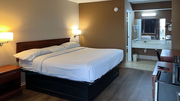 A hotel room with a bed, bedside lamps, a wooden nightstand, a desk with a chair, a mirror, and a bathroom with a sink and mirror. - Studio 6 Millington, Tn (Millington)