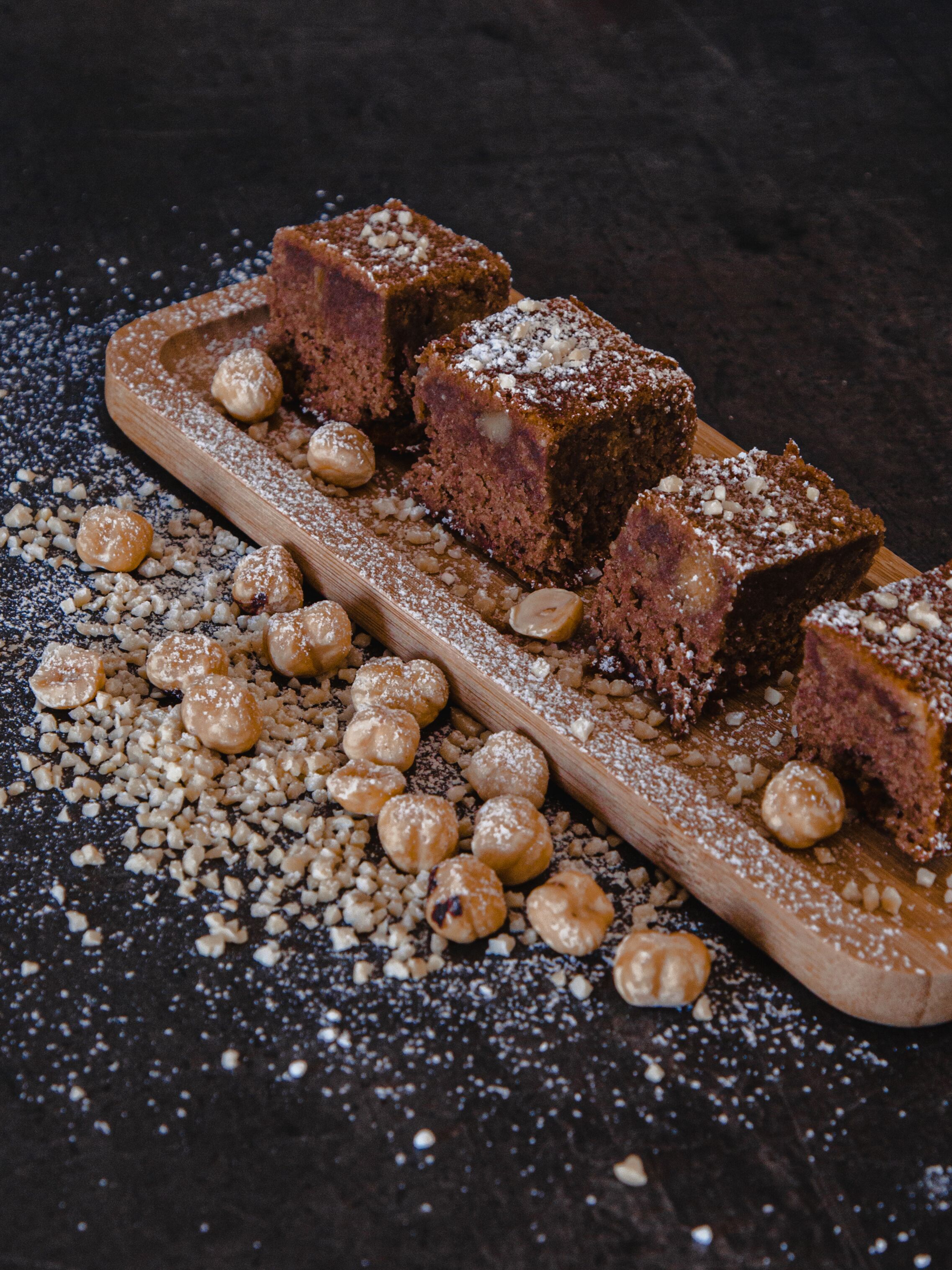 A wooden serving board with several square pieces of cake and hazelnuts scattered around.