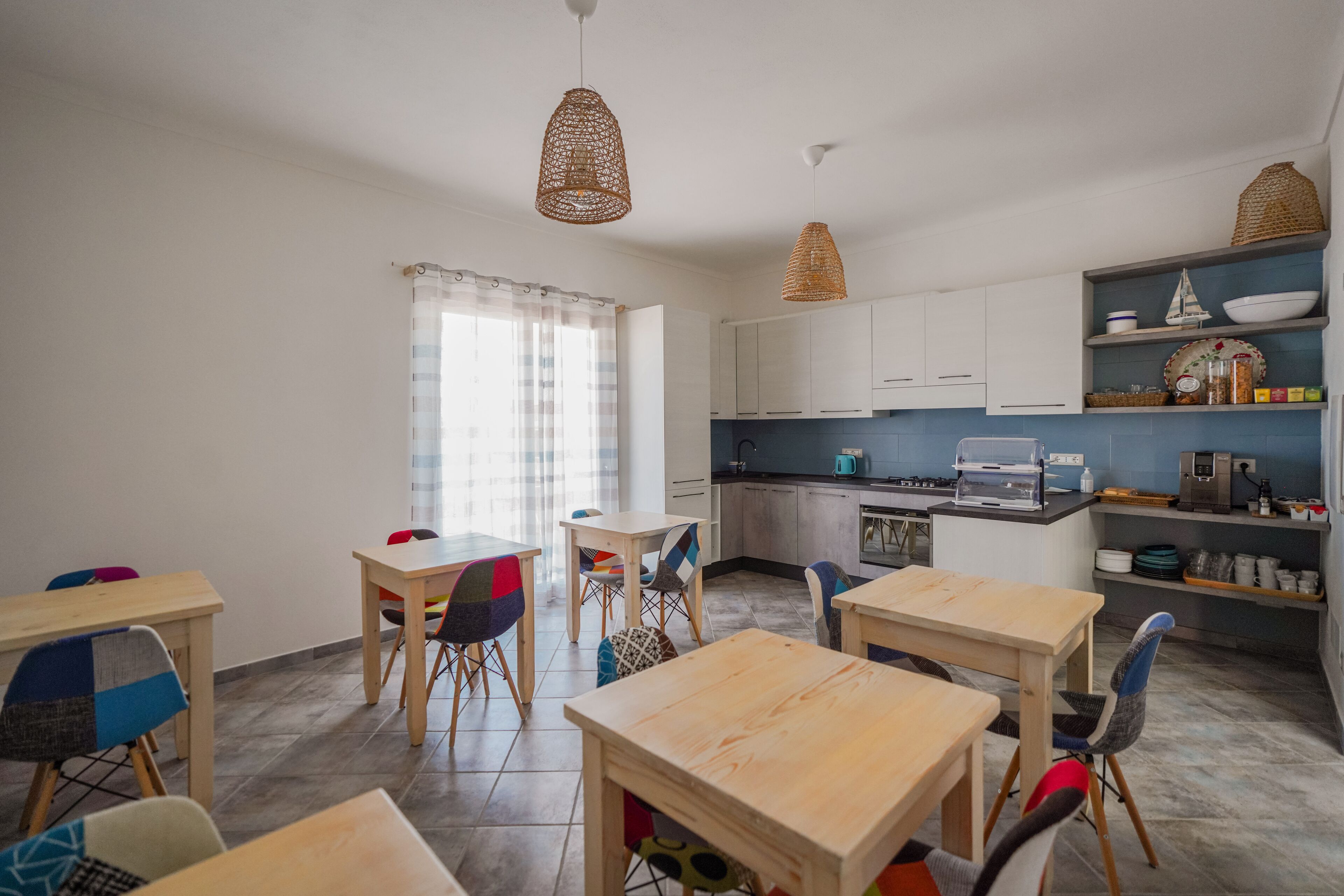 A modern kitchen with white cabinets, a dining area with wooden tables and colorful chairs, and a window with sheer curtains.