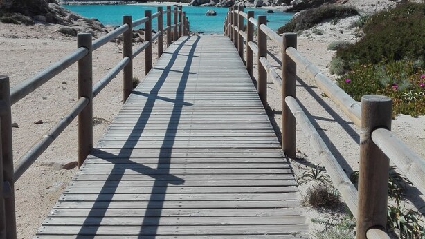 Una pasarela de madera que lleva a una playa con agua turquesa cristalina y colinas rocosas al fondo.