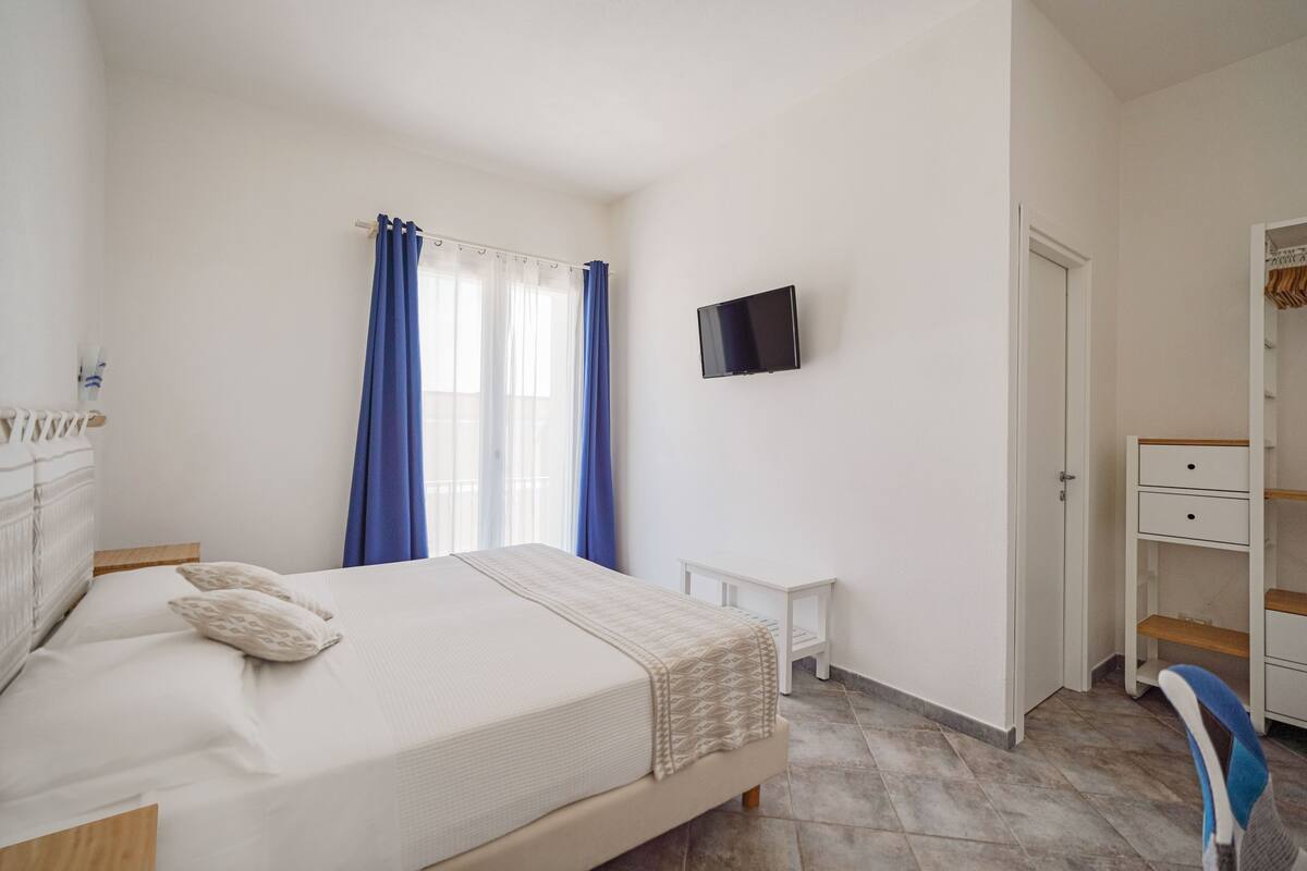Un dormitorio con cama, televisor montado en la pared, una mesita de noche pequeña y un armario.