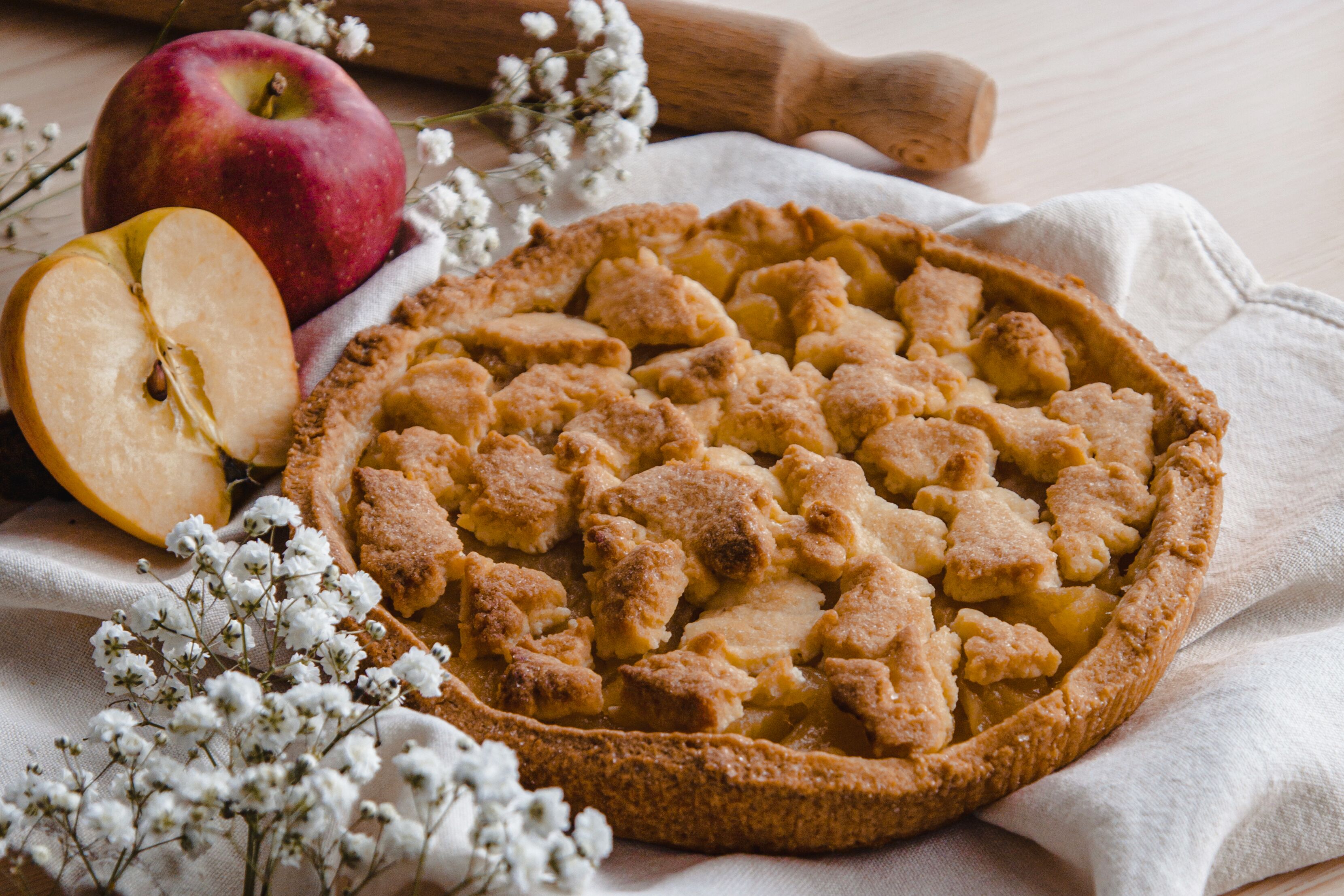 Una tarta de manzana con tapa de rejilla, rodeada de un paño, una manzana y algunas flores.