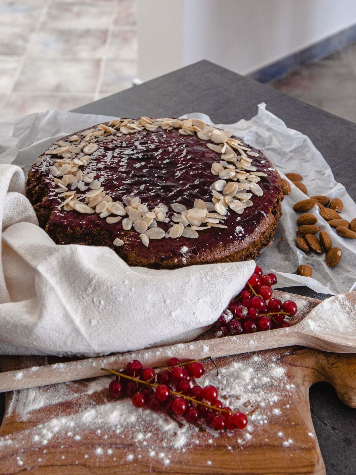 A round cake with a glossy topping, almonds, and red berries on a wooden board.