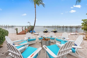 A beachside seating area with white chairs and a fire pit, overlooking a calm sea and a pier. - Aquarius South: Luxe 10BR/6BA , w/ heated swimming pool+spa, private dock & more (Stuart)