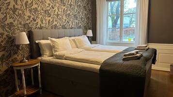 Cama doble con cabecera gris y ropa de cama blanca, una mesita de noche con lámpara y una ventana con cortinas.
