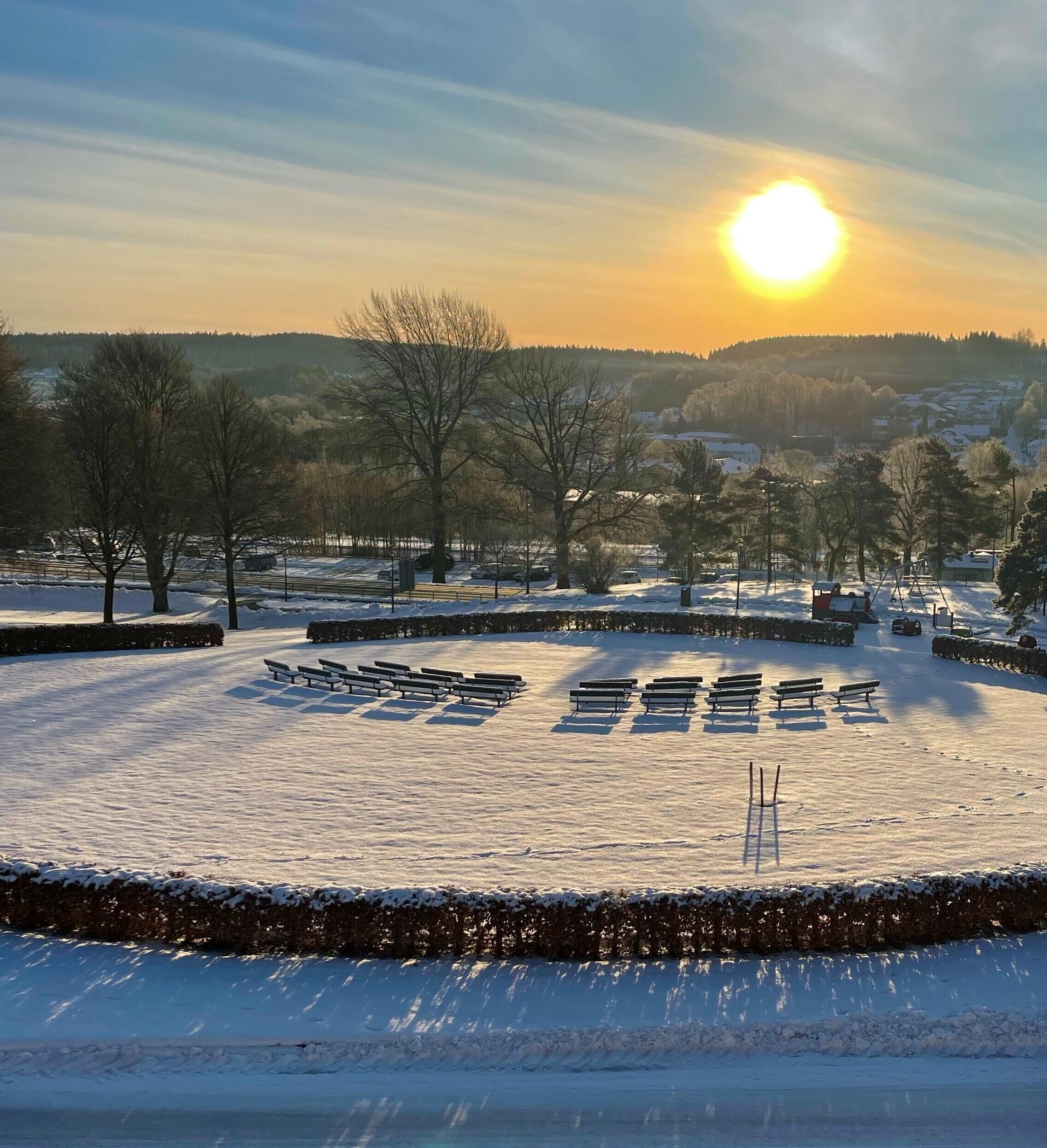 A snow-covered park with benches and trees, a clear sky, and a setting sun.