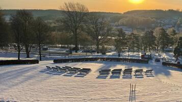 A snow-covered park with benches and trees, a clear sky, and a setting sun.