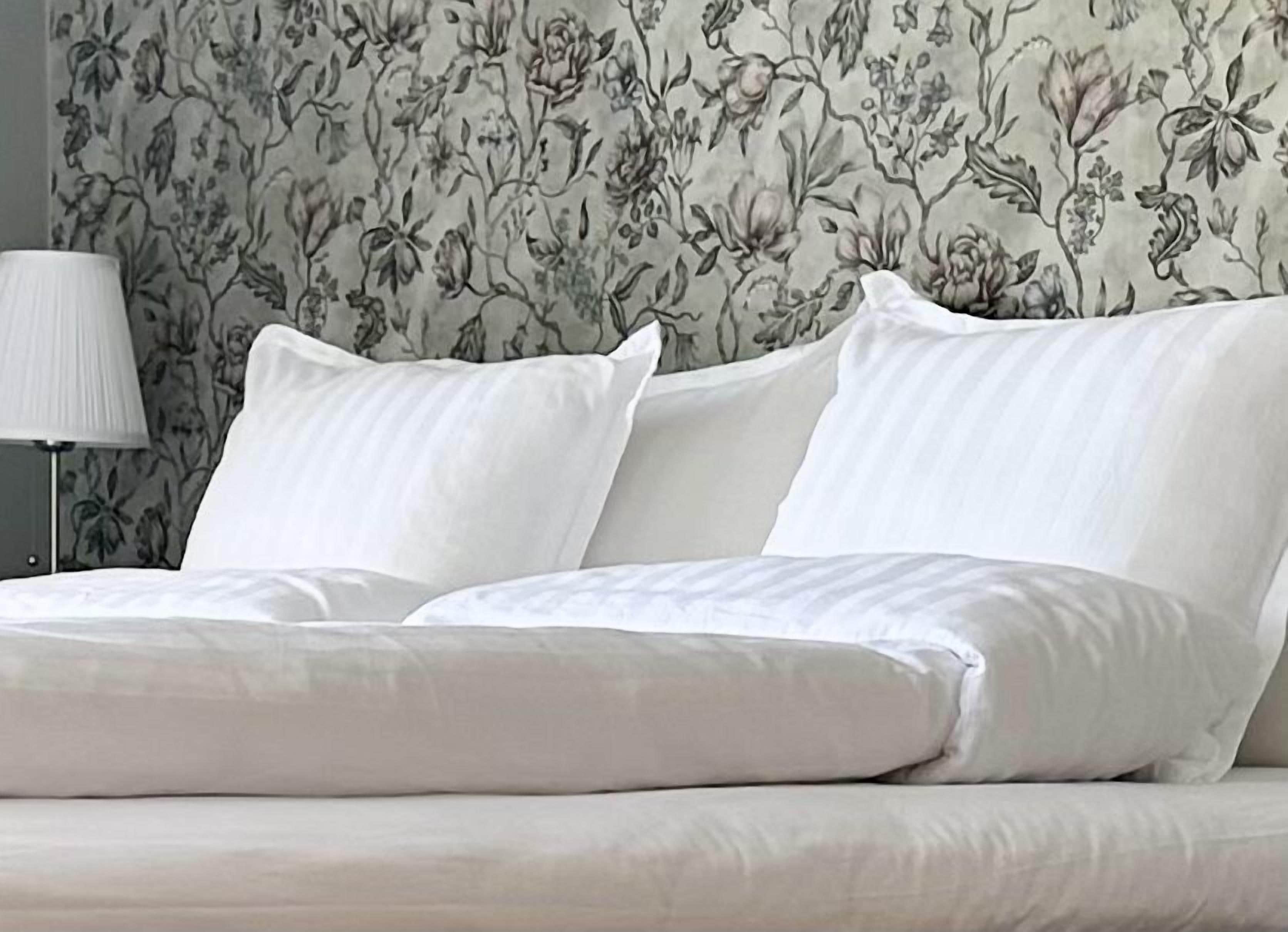 Un letto con lenzuola e cuscini bianchi, un comodino con una lampada e una carta da parati con motivo floreale.