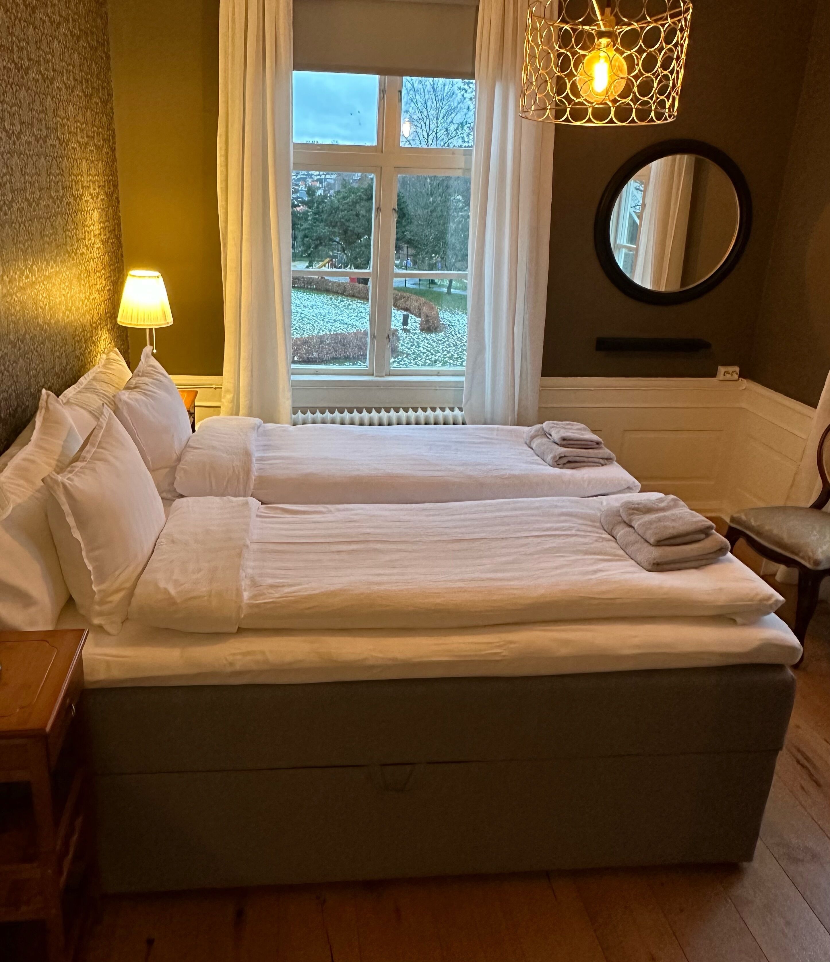 A double bed with white linens, a bedside lamp, a round mirror, and a view of a park through a window.