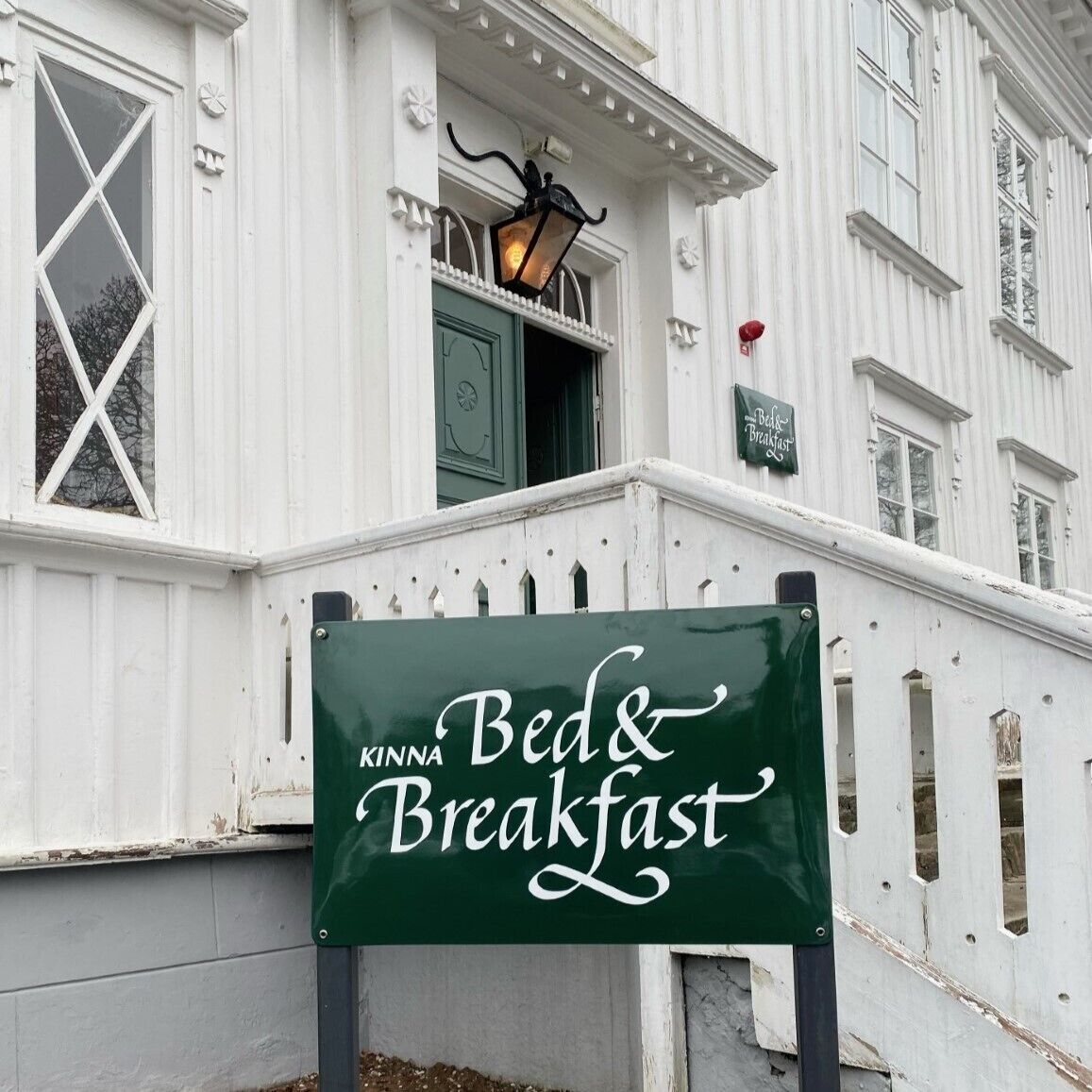Skilt med 'Kinna Bed & Breakfast' foran en hvid bygning med en grøn dør og en lygt over døren.