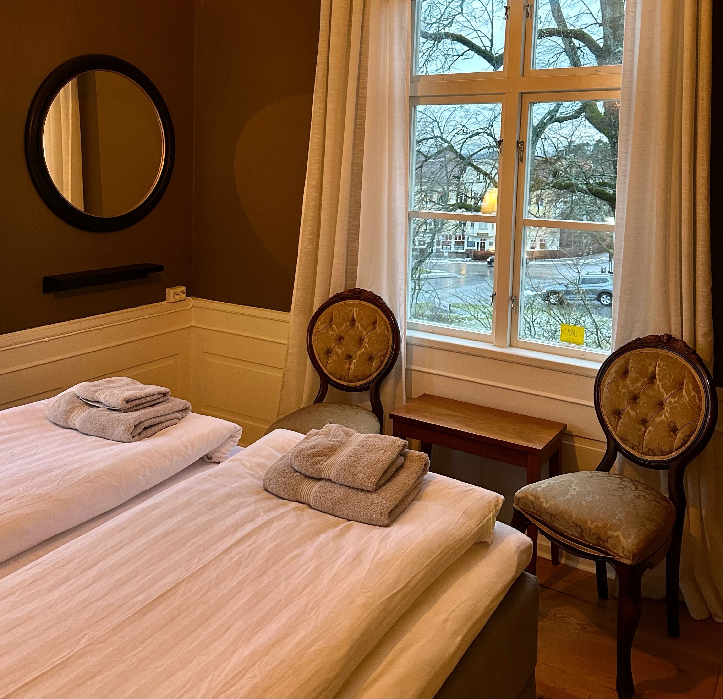 Ein Hotelzimmer mit zwei Betten, einem Spiegel, einem Fenster mit Außenblick und zwei Polsterstühlen.
