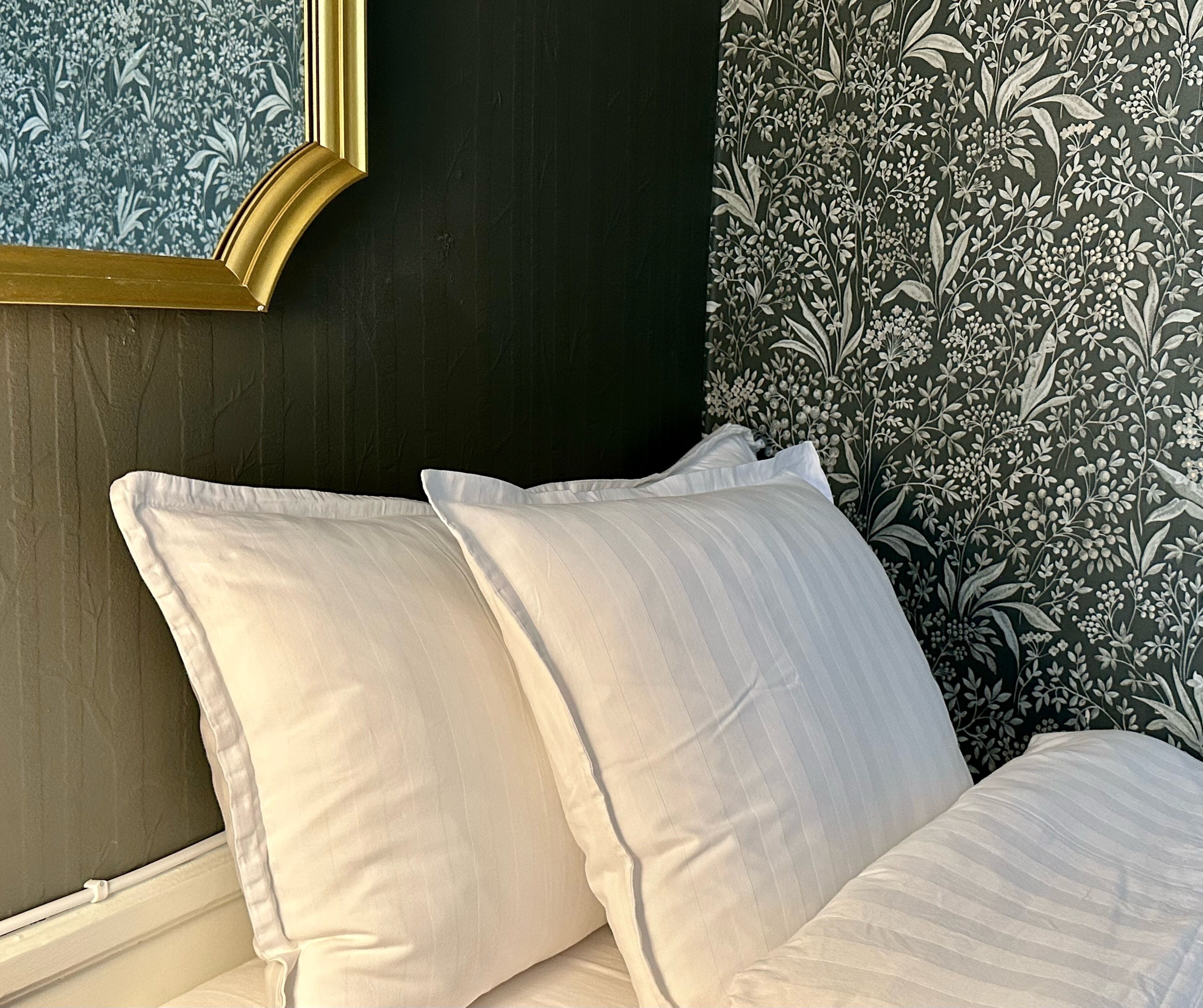 Una cama con almohadas blancas y una pared oscura con papel tapiz floral.