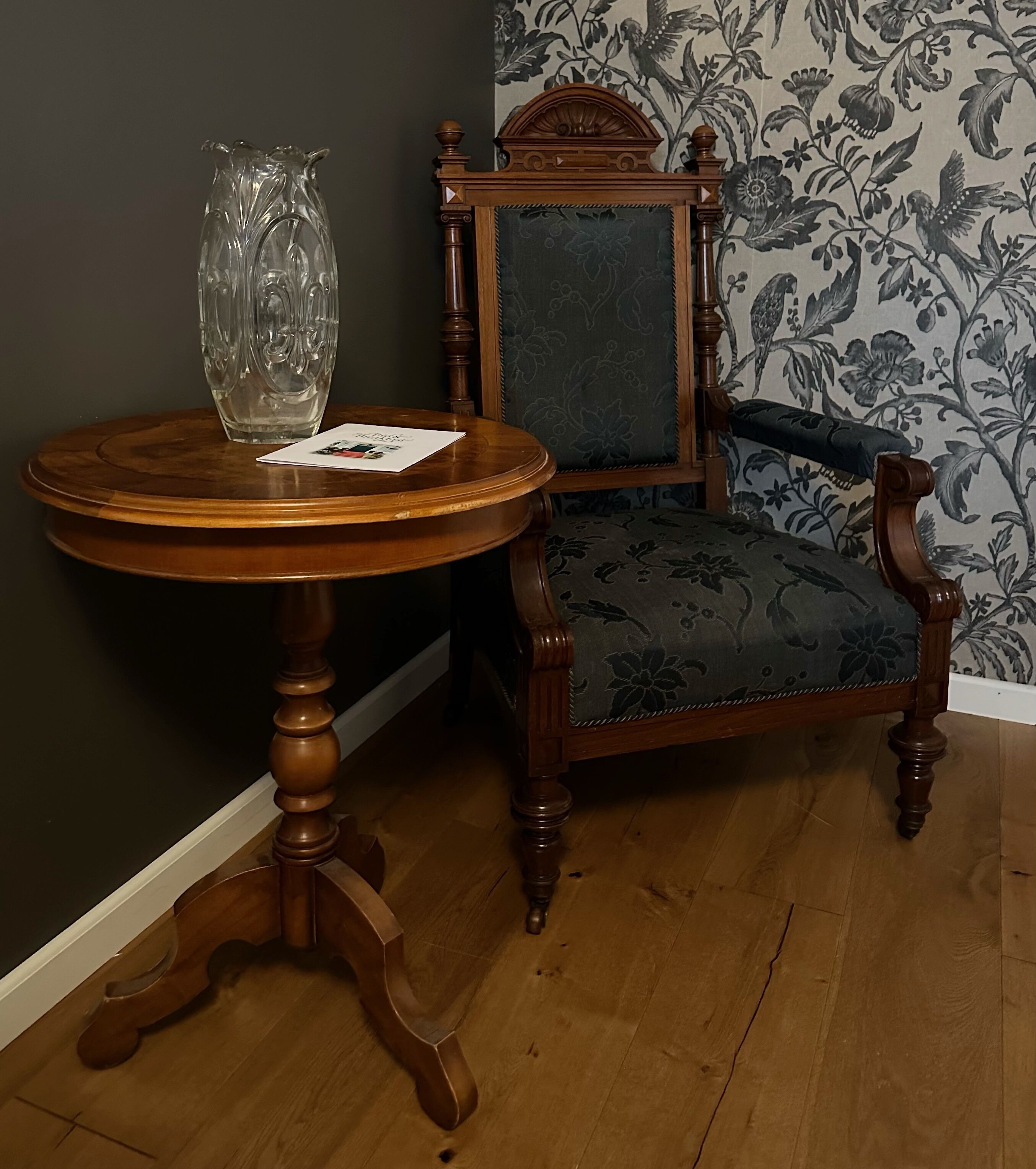 A wooden table with a vase and a card, a chair with a floral upholstery, and a floral wallpaper pattern.