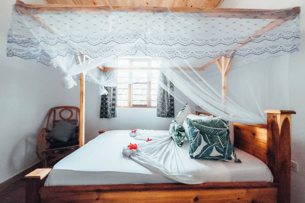 A wooden bed with a canopy, a window with patterned curtains, and a chair with a cushion.