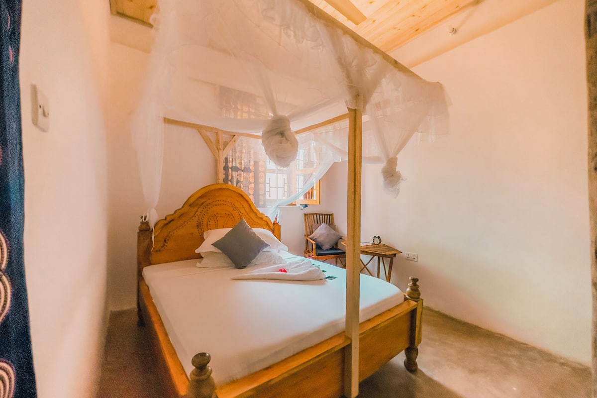 A four-poster canopy bed with a wooden headboard, a bedside table, and a chair.