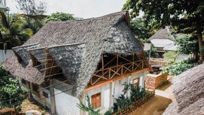 Une maison à toit de chaume, dotée d’un balcon en bois, entourée de verdure et bordée d’un chemin de terre.