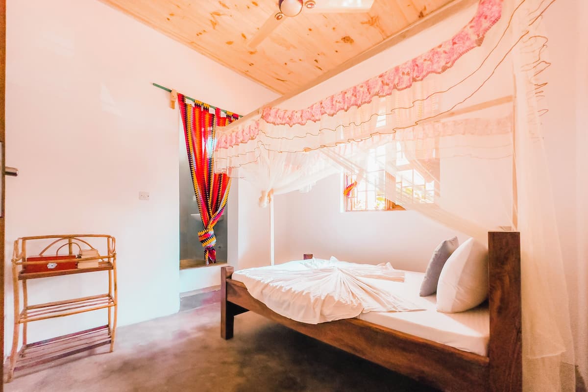 A bedroom with a wooden bed, a bedside table, a window with sheer curtains, and a colorful hanging decoration.