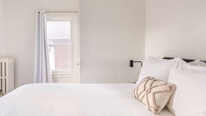 Une chambre avec un grand lit, du linge de lit blanc, un radiateur, une fenêtre avec des rideaux et un mur de briques visible à travers la fenêtre.