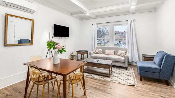 Un salon moderne avec une table à manger en bois, des chaises transparentes, un canapé et un fauteuil bleu. Un téléviseur à écran plat est fixé au mur, et il y a une fenêtre donnant sur la ville.
