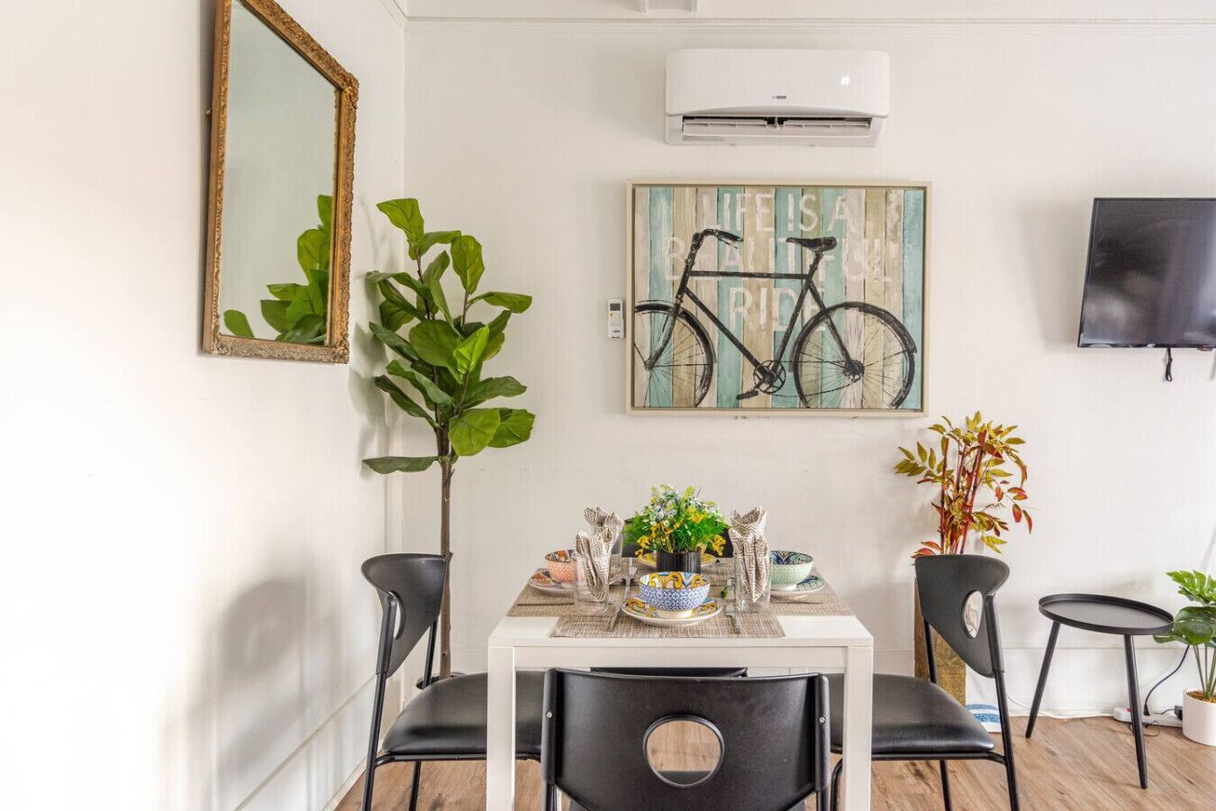 A dining area with a white table, black chairs, a mirror, an air conditioner, and decorative items including a bicycle painting and plants.